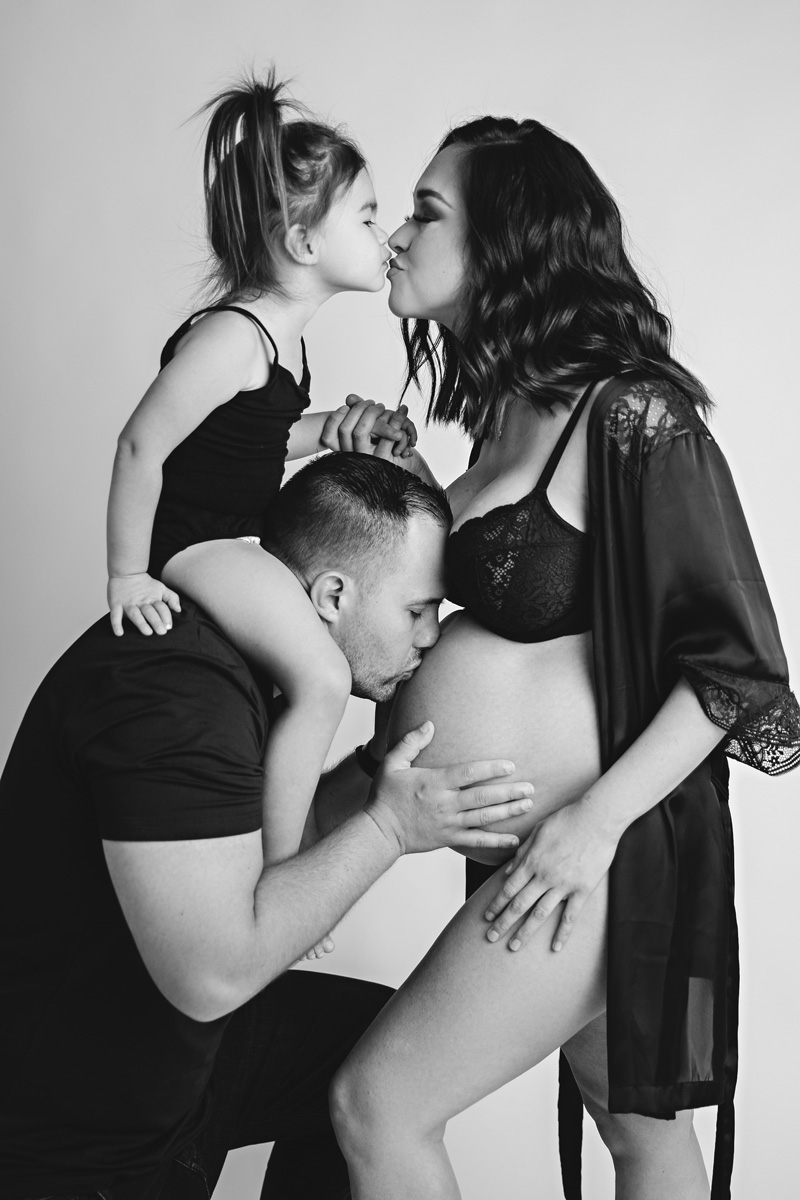 Boise modern maternity — black-and-white family portrait, partner kissing the belly while toddler kisses mother
